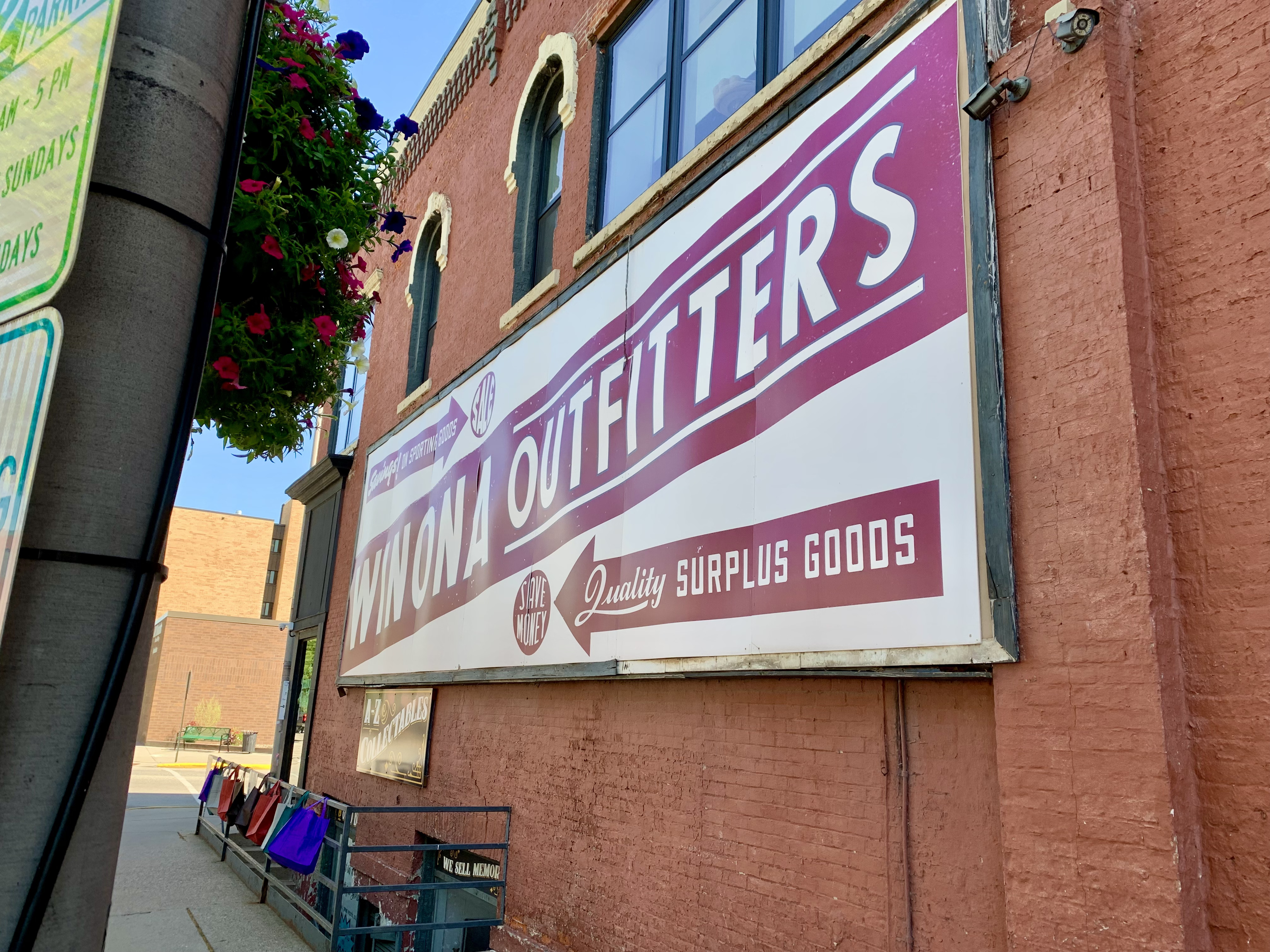 A billboard with a white background, a brown stripe and arrows, and white text on those brown elements. The billboard reads Winona Outfitters and quality surplus goods. The billboard is in a wooden frame on the side of a reddish-brown brick building.